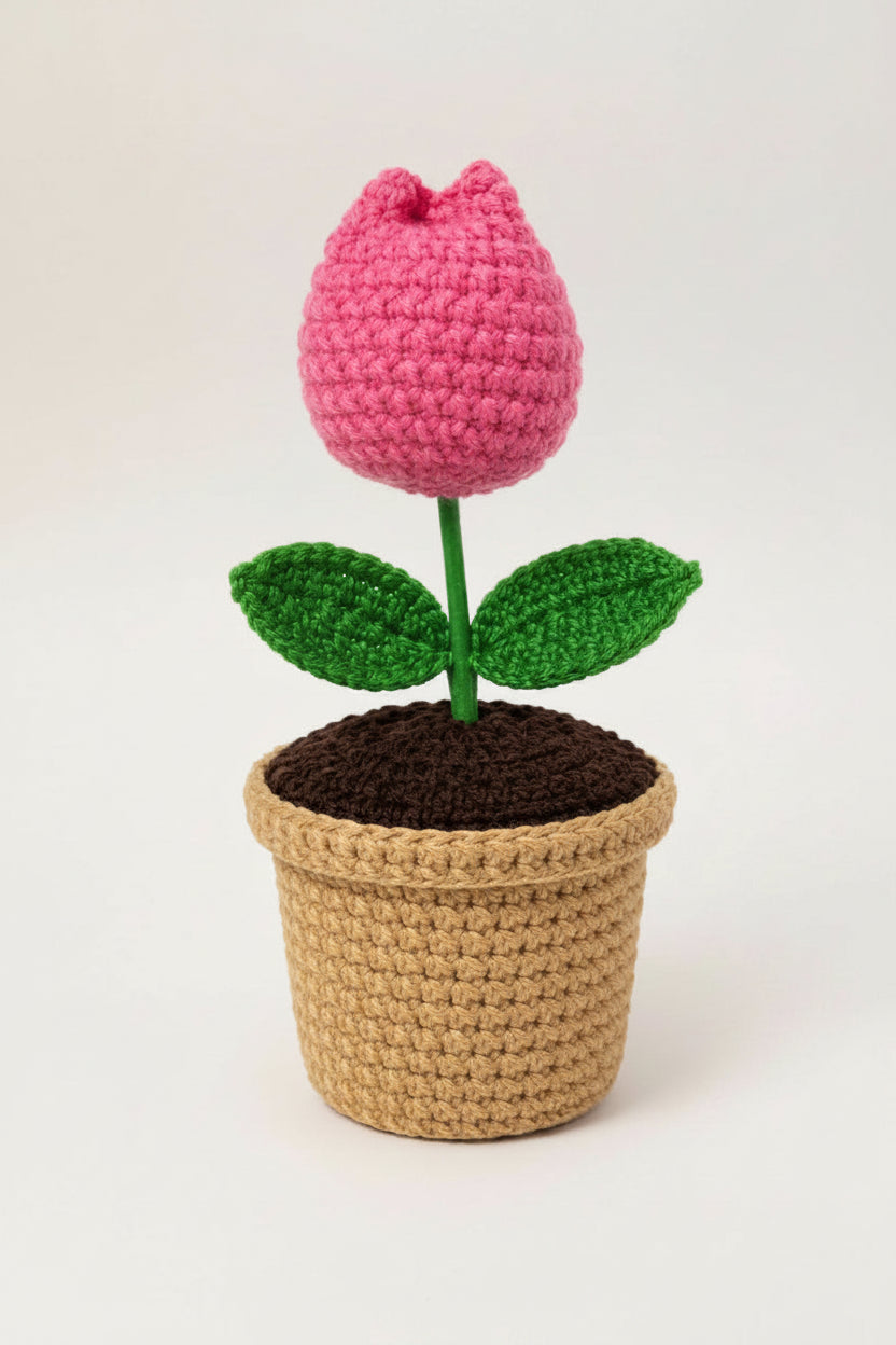 A crocheted flower pot plant with a pink flower head, green stem and leaves, and a beige woven pot on a white background.
