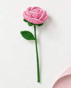 A crocheted pink rose with a green stem and leaf, photographed on a clean white background with a soft pink paper corner visible in the lower right.