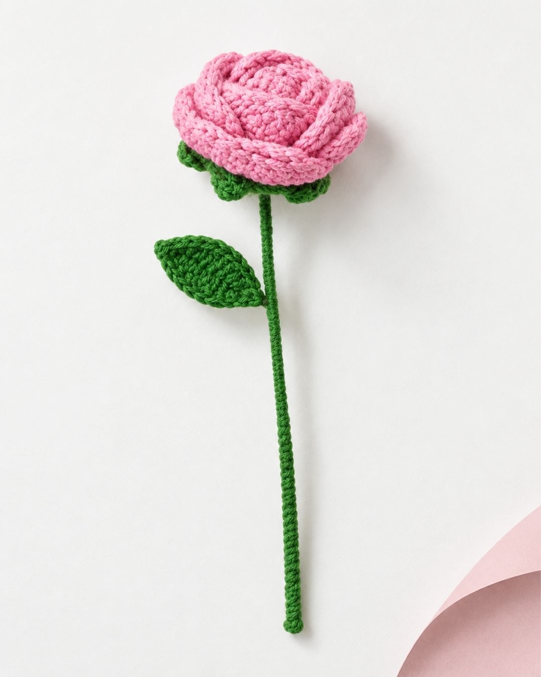 A crocheted pink rose with a green stem and leaf, photographed on a clean white background with a soft pink paper corner visible in the lower right.