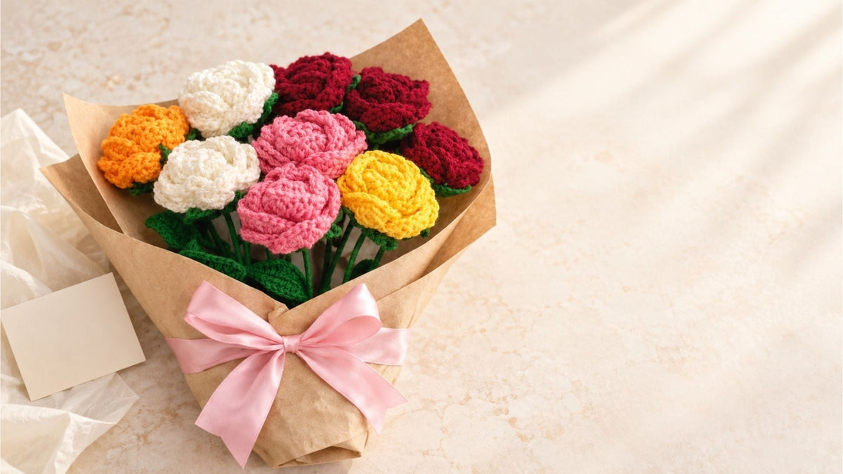 A bouquet of crocheted flowers wrapped in brown kraft paper and tied with a pink ribbon, placed on a light beige surface. The flowers include red, pink, yellow, white, and orange, with green stems visible at the bottom.