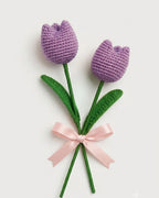 Two crocheted tulip flowers with purple blooms, green stems, green leaves, and a light pink ribbon tied around the stems, shown on a plain light background.