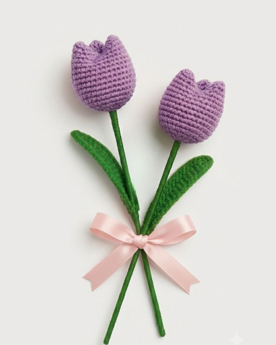 Two crocheted tulip flowers with purple blooms, green stems, green leaves, and a light pink ribbon tied around the stems, shown on a plain light background.