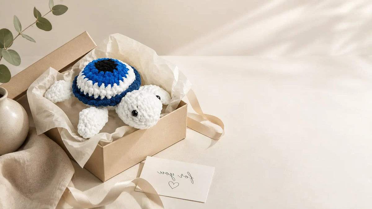 A crocheted white turtle toy wearing a blue and white striped hat, displayed inside a kraft gift box with tissue paper and a small card that reads 'for you'.